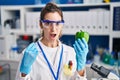 Young hispanic woman working at scientist laboratory with vegetables in shock face, looking skeptical and sarcastic, surprised