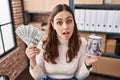 Young hispanic woman working at small business ecommerce holding money and piggy bank in shock face, looking skeptical and Royalty Free Stock Photo