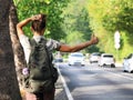 Young hitchhiker with a backpack behind her, looks towards the approaching car