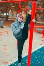 Young hooded gymnast woman warming up at playground pillars and doing splits in park at vibrant autumn