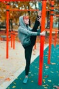 Young hooded parkour woman warming up at playground pillars and doing splits in park at autumn