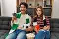 Young hooligan couple supporting soccer team sitting on the sofa at home