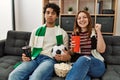 Young hooligan couple supporting soccer team sitting on the sofa at home