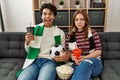 Young hooligan couple supporting soccer team sitting on the sofa at home