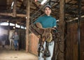 Young horsewoman walking in stable with saddle in hands
