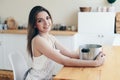 Woman in an Apron Smiling sits at wooden Table. Blurred Kitchen in Background. Think What to Cook