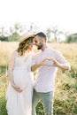 Young husband and his pregnant wife in hat and white dress, walking in the sunny summer field. Man makes a heart gesture Royalty Free Stock Photo