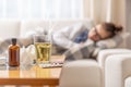 Young ill woman lying under blanket on a bed with seasonal illnes drinking hot tea with honey and lemon. She takes pills and cough