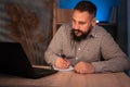 Young indian businessman using a laptop and writing notes while working overtime at his home office desk late in the Royalty Free Stock Photo