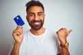 Young indian customer man holding credit card standing over isolated white background pointing and showing with thumb up to the Royalty Free Stock Photo