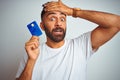 Young indian customer man holding credit card standing over isolated white background stressed with hand on head, shocked with Royalty Free Stock Photo