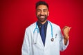 Young indian doctor man wearing stethoscope standing over isolated red background smiling with happy face looking and pointing to Royalty Free Stock Photo