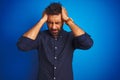 Young indian elegant man wearing shirt standing over isolated blue background suffering from headache desperate and stressed