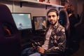 A young Indian guy wearing a military shirt sitting on a gamer chair and looking at a camera in a gaming club or