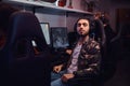 A young Indian guy wearing a military shirt sitting on a gamer chair and looking at a camera in a gaming club or