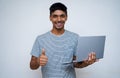 `Young indian handsome boy showing thumbs up and smiling while looking into the camera, holding laptop in his other hand Royalty Free Stock Photo