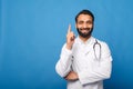 Young indian male medical worker wearing white gown with stethoscope over shoulders standing isolated blue