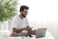 Young Indian Man Sitting In Bed Using Laptop And Writing In Notepad Royalty Free Stock Photo