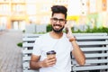 Young freelancer indian man sitting on bench, talking by phone, looking at camera. Handsome man in glasses wearing in white Royalty Free Stock Photo