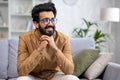 A young Indian man is sitting smiling on the sofa at home, holding his hands together and resting his chin on them Royalty Free Stock Photo