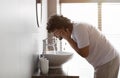 Young indian man washing face with clear water from tap bending over sink, doing skincare facial routine in bathroom Royalty Free Stock Photo