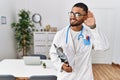 Young indian man wearing doctor uniform and stethoscope smiling with hand over ear listening an hearing to rumor or gossip Royalty Free Stock Photo