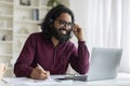 Young indian man wearing headset using laptop and smiling while taking notes Royalty Free Stock Photo