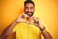 Young indian man wearing polo standing over isolated yellow background smiling in love showing heart symbol and shape with hands Royalty Free Stock Photo