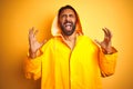 Young indian man wearing raincoat with hood standing over isolated yellow background celebrating mad and crazy for success with
