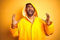 Young indian man wearing raincoat with hood standing over isolated yellow background crazy and mad shouting and yelling with