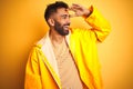 Young indian man wearing raincoat standing over isolated yellow background very happy and smiling looking far away with hand over Royalty Free Stock Photo