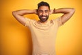 Young indian man wearing t-shirt standing over isolated yellow background relaxing and stretching, arms and hands behind head and Royalty Free Stock Photo