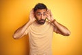 Young indian man wearing t-shirt standing over isolated yellow background Trying to hear both hands on ear gesture, curious for