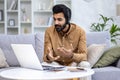 A young Indian man works remotely from home. He sits on the couch in a headset in front of a laptop and chats on a video Royalty Free Stock Photo