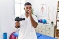 Young indian physiotherapist holding therapy massage gun at wellness center looking stressed and nervous with hands on mouth Royalty Free Stock Photo