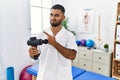 Young indian physiotherapist holding therapy massage gun at wellness center pointing aside worried and nervous with forefinger, Royalty Free Stock Photo