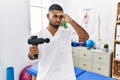 Young indian physiotherapist holding therapy massage gun at wellness center worried and stressed about a problem with hand on Royalty Free Stock Photo