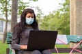 Young Indian woman with mask using laptop while sitting with distance on park bench Royalty Free Stock Photo