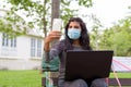 Young Indian woman with mask video calling using phone while sitting with distance on park bench Royalty Free Stock Photo