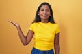 Young indian woman standing over yellow background smiling cheerful presenting and pointing with palm of hand looking at the Royalty Free Stock Photo