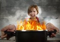 Young inexperienced home cook woman in panic with apron holding pot burning in flames with in panic