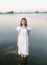 a young innocent girl in a light dress with a bow is standing in the river at dawn