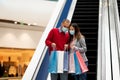 Young international couple in protective masks going down on escalator at mall, looking inside shopping bags, free space Royalty Free Stock Photo