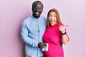 Young interracial couple expecting a baby holding shoes pointing thumb up to the side smiling happy with open mouth Royalty Free Stock Photo