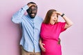 Young interracial couple expecting a baby, touching pregnant belly very happy and smiling looking far away with hand over head Royalty Free Stock Photo