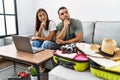 Young interracial couple packing summer clothes in suitcase looking at laptop serious face thinking about question with hand on Royalty Free Stock Photo