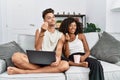 Young interracial couple using laptop at home sitting on the sofa doing money gesture with hands, asking for salary payment, Royalty Free Stock Photo