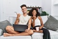 Young interracial couple using laptop at home sitting on the sofa pointing to the back behind with hand and thumbs up, smiling Royalty Free Stock Photo