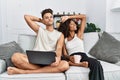 Young interracial couple using laptop at home sitting on the sofa smiling confident touching hair with hand up gesture, posing Royalty Free Stock Photo