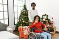 Young interracial couple with woman sitting on wheelchair by christmas tree showing and pointing up with fingers number four while